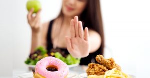 an image of a woman pushing away donuts and fried chicken with her left hand as she holds a green apple in her right hand to signify the importance of a healthy diet